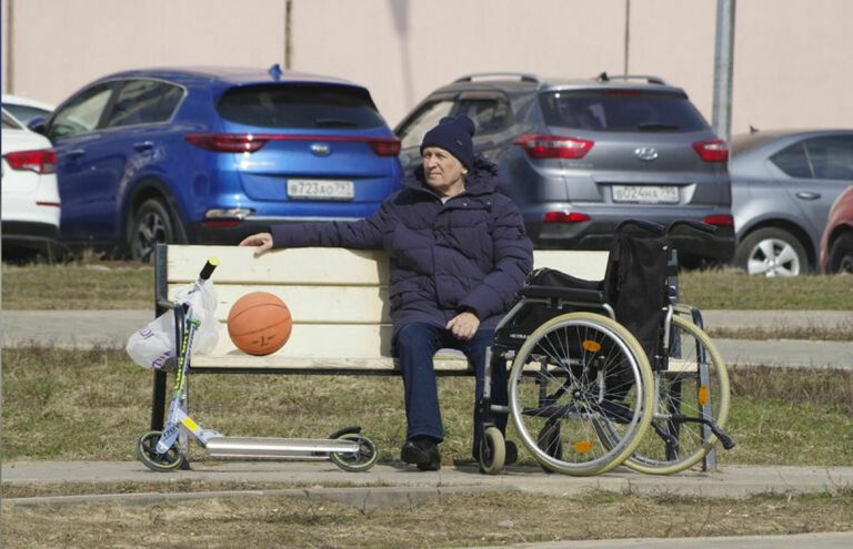 цветная фотография. Солнечный день. На белой лавочке сидит пожилой мужчина в теплой синей куртке и шапке. Рядом с ним стоят кресло-коляска и самокат, на скамейке лежит баскетбольный мяч. Позади мужчины плотно заполненная автомобильная стоянка.