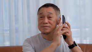 Мужчина в серой футболке поднес телефон к уху, сидя на коричневом диване на фоне полупрозрачных занавесок.