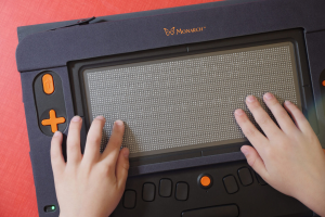 Тактильный дисплей для слабовидящих и незрячих людей марки Monarch. На экране расположены рельефные точки Брайля, по которым проводят руками. Слева видны оранжевые кнопки управления, внизу — панель с клавишами.